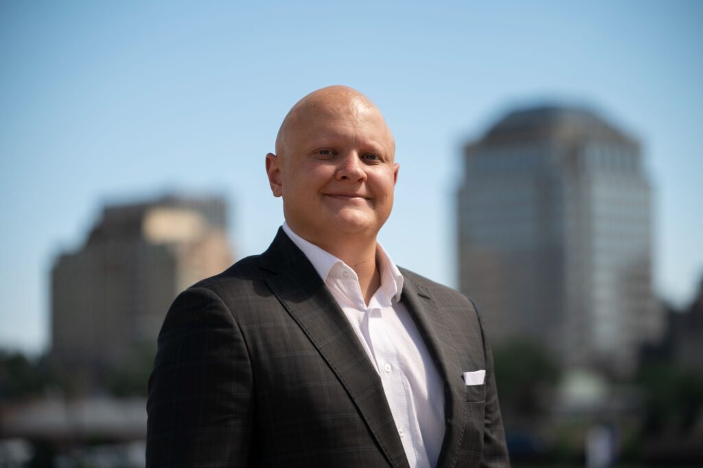 A bald man in a suit standing in front of a city.