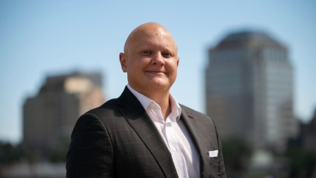 A bald man in a suit standing in front of a city.