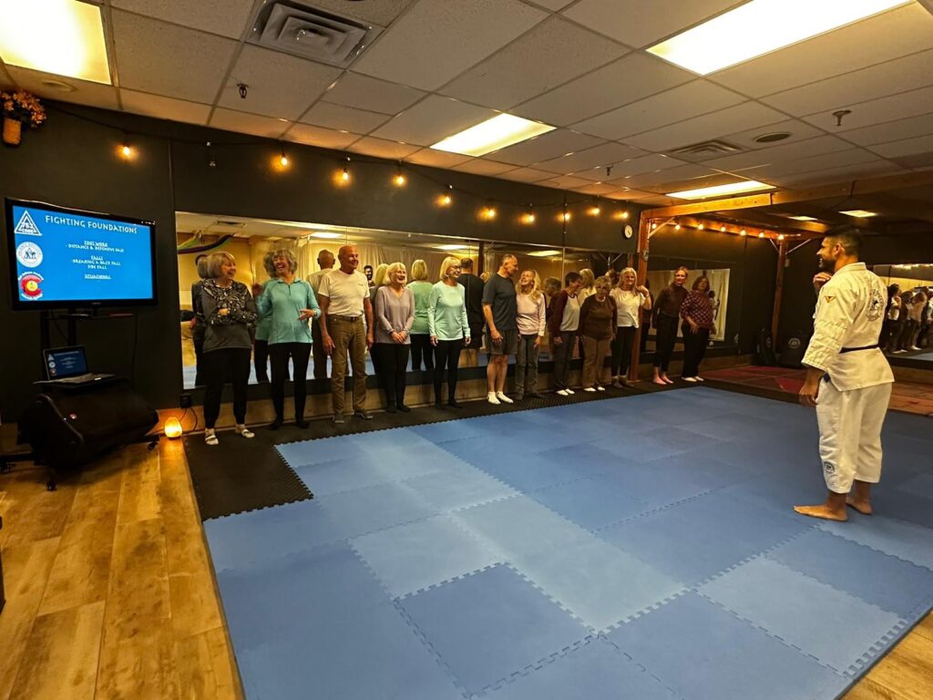 A group of adults stands on a blue mat facing a man in a white gi in a martial arts dojo, attentive to a presentation displayed on a screen.