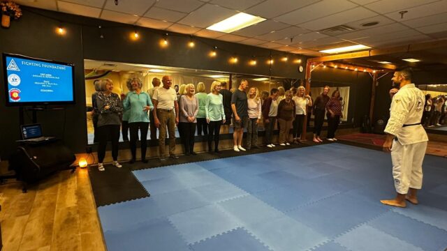 A group of adults stands on a blue mat facing a man in a white gi in a martial arts dojo, attentive to a presentation displayed on a screen.