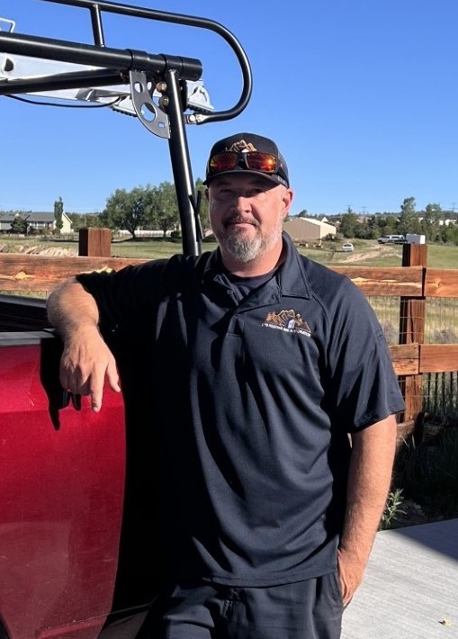 Middle-aged man with sunglasses, beard, and black polo shirt standing by a red vehicle, with a wooden fence and grassy field in the background.