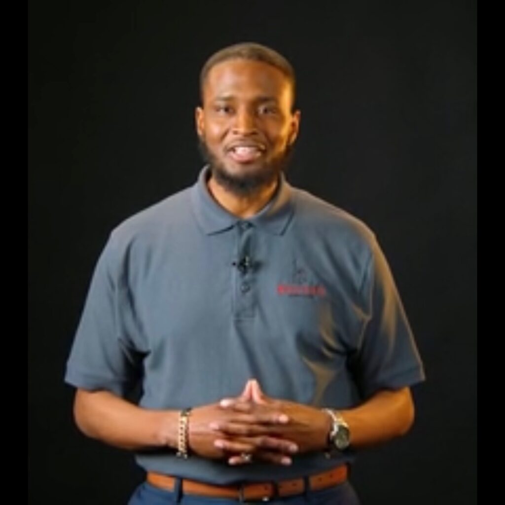 A man wearing a grey polo shirt with a logo stands with his hands clasped, posing against a plain black background.