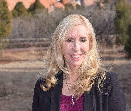 A woman with long blonde hair, wearing a black blazer and purple top, stands outdoors with a rocky landscape and trees in the background.