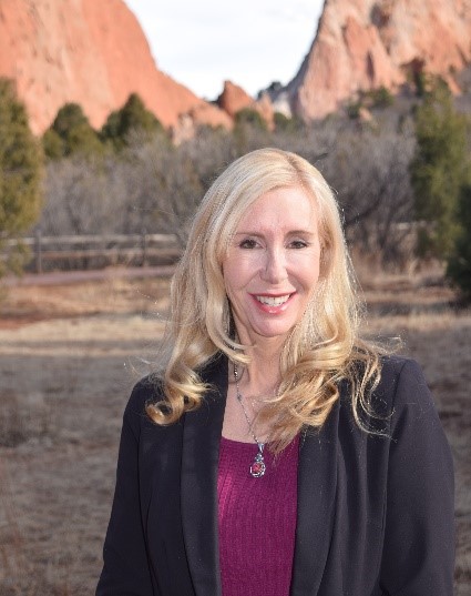 A woman with long blonde hair, wearing a black blazer and purple top, stands outdoors with a rocky landscape and trees in the background.