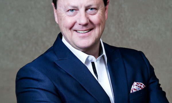 Smiling man in a navy blazer, white shirt, and pocket square stands with arms crossed against a textured brown background.