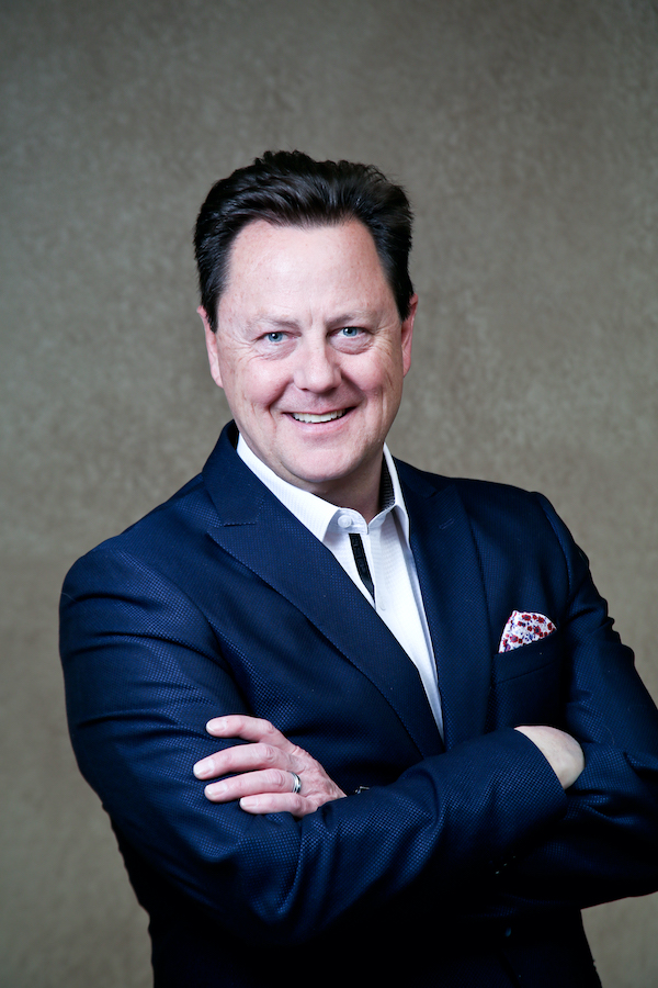 Smiling man in a navy blazer, white shirt, and pocket square stands with arms crossed against a textured brown background.