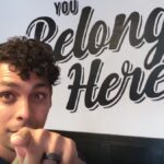 Person points towards the camera in front of a wall with the text "You Belong Here.