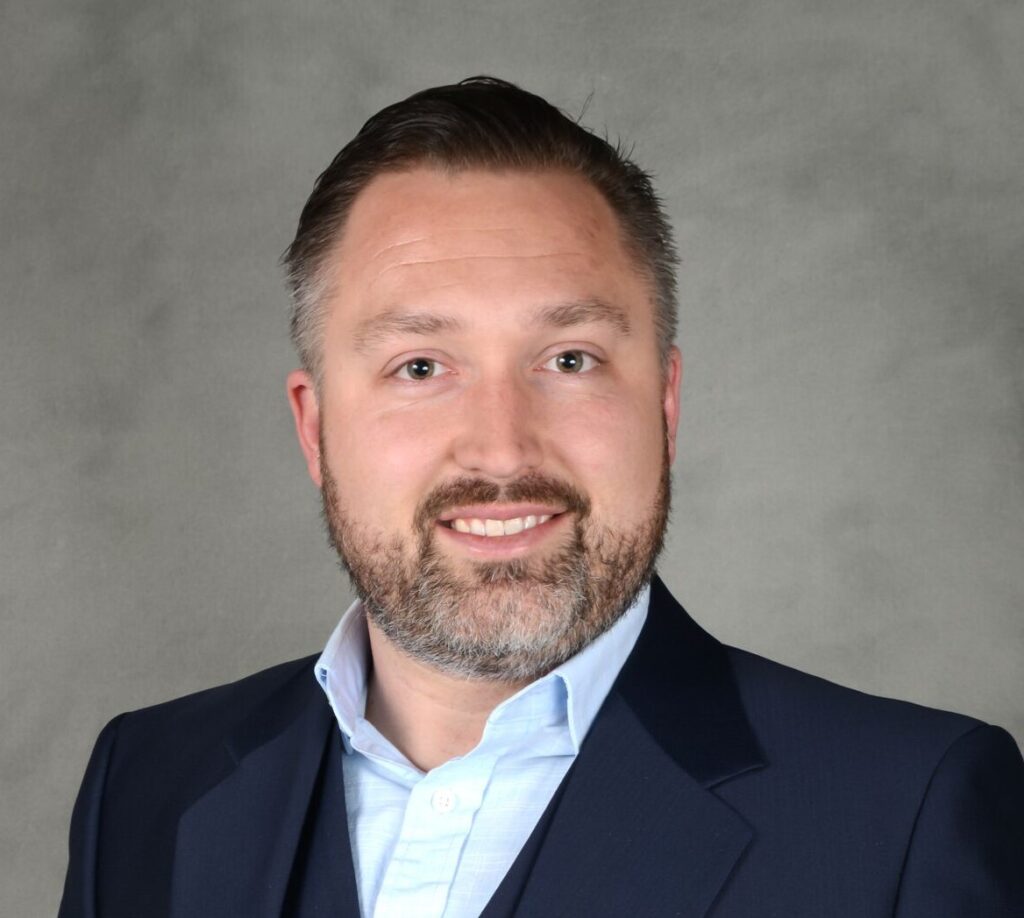 A man with a beard and short hair is wearing a suit and blue shirt, smiling against a gray background.