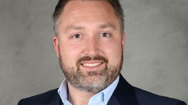 A man with a beard and short hair is wearing a suit and blue shirt, smiling against a gray background.