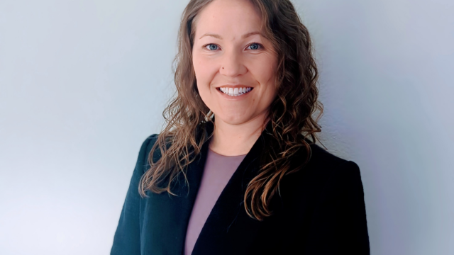 Person with long curly hair, wearing a dark blazer, smiling against a plain light background.