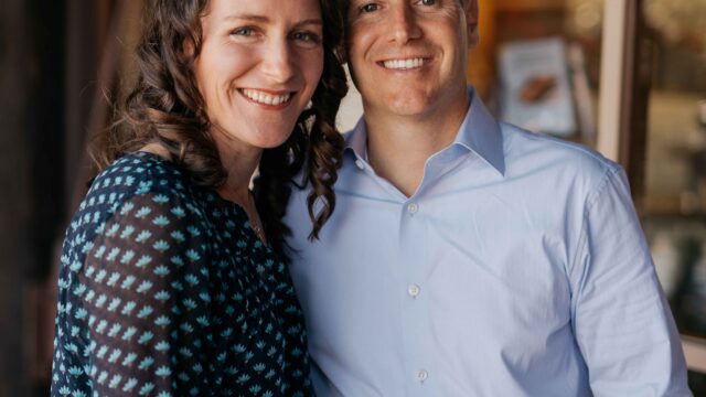 A woman and a man stand closely together, smiling, in a casual setting near large windows.