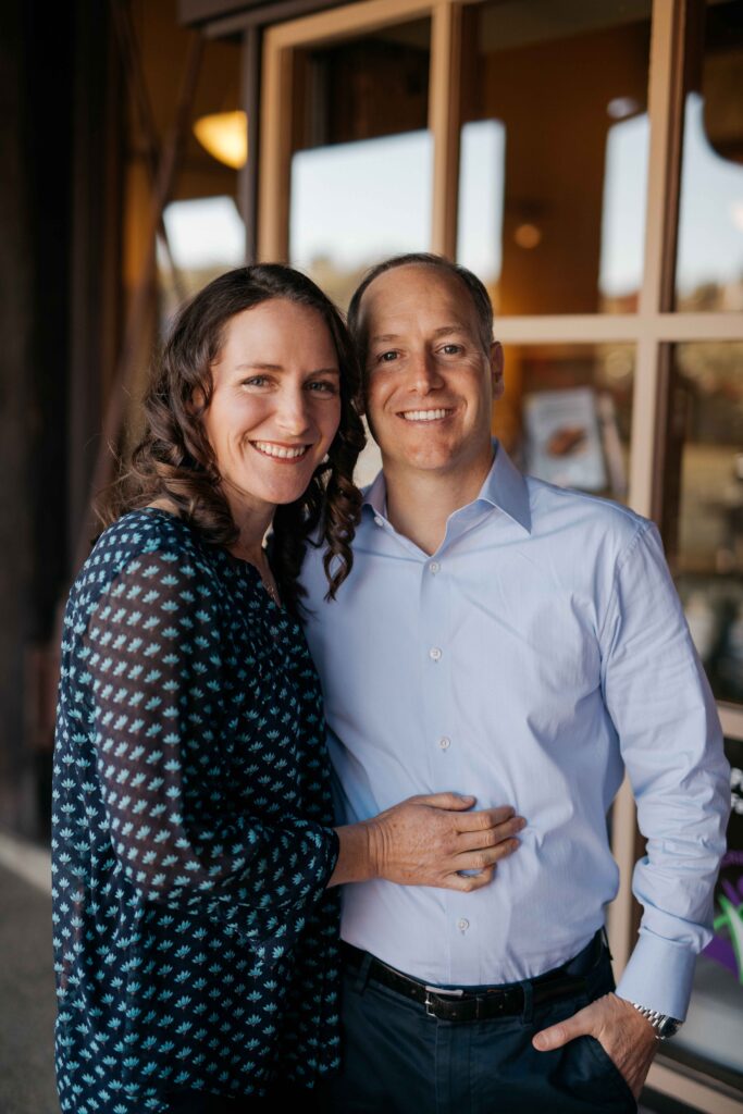 A woman and a man stand closely together, smiling, in a casual setting near large windows.