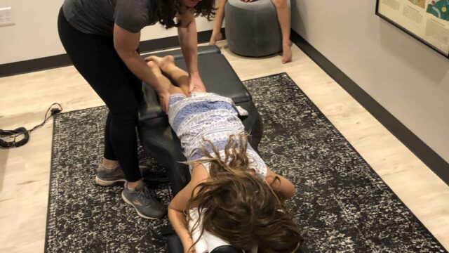 A chiropractor adjusts a child's back on a treatment table while another child sits nearby in an office setting.