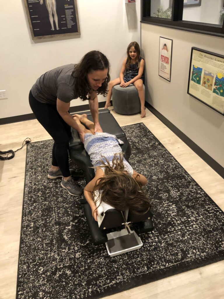 A chiropractor adjusts a child's back on a treatment table while another child sits nearby in an office setting.