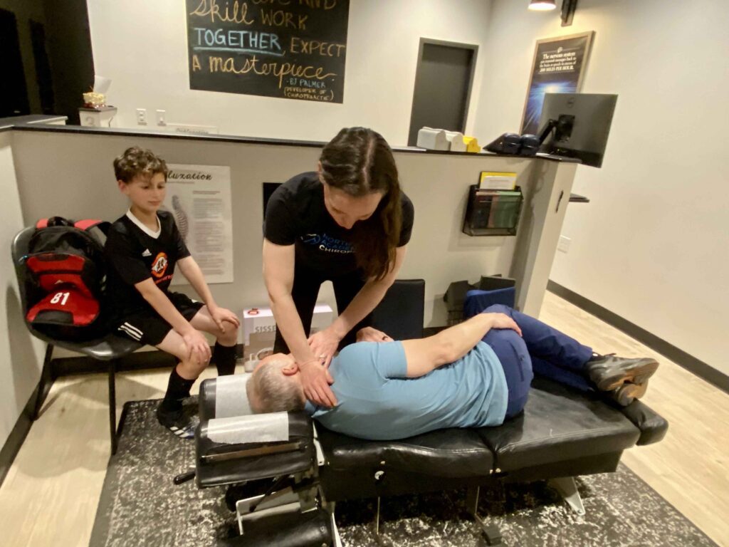 A chiropractor adjusts a man's neck on a treatment table, while a seated boy watches nearby in a room with a motivational wall sign.