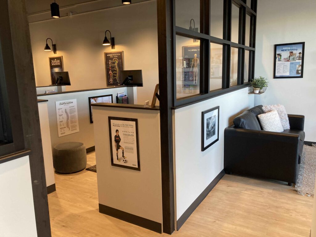 Chiropractic office with a waiting area featuring a black couch and decorative items, alongside individual consultation spaces divided by partitions. Posters and diagrams adorn the walls.