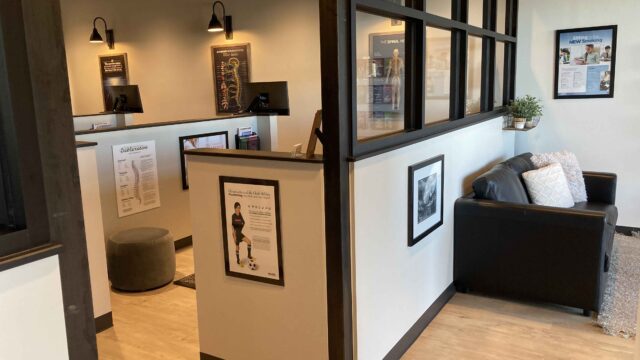 Chiropractic office with a waiting area featuring a black couch and decorative items, alongside individual consultation spaces divided by partitions. Posters and diagrams adorn the walls.