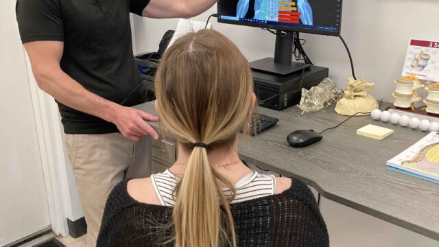 A person in a black shirt discusses a spinal diagram on a monitor with a seated woman in a consultation room.