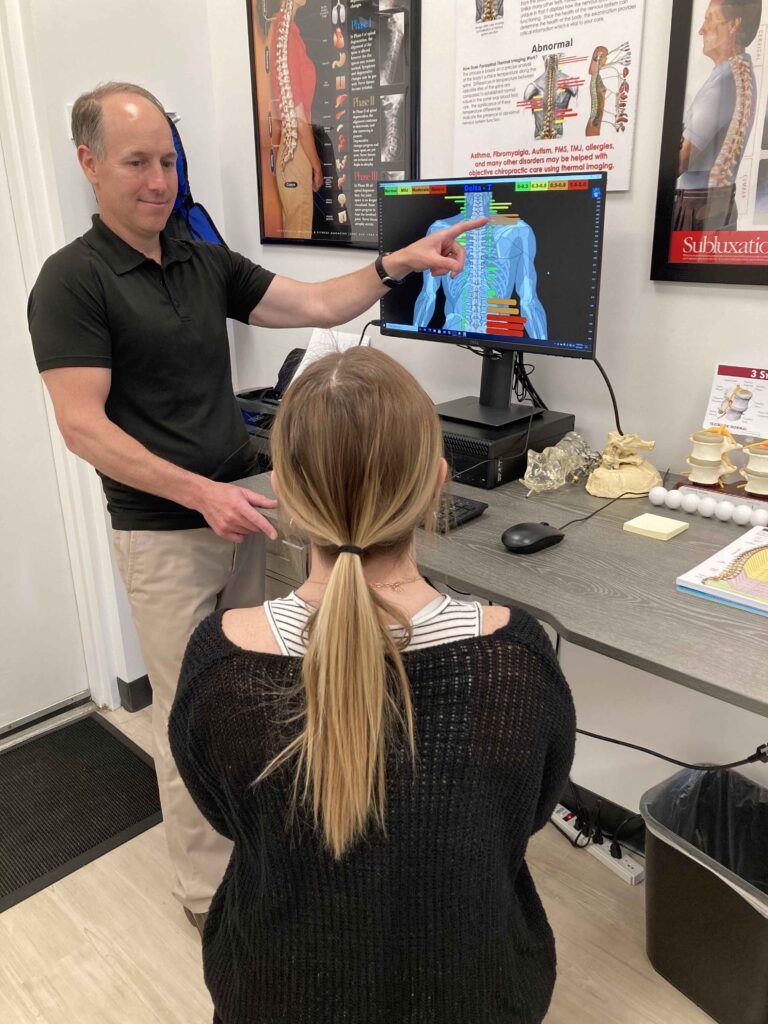 A person in a black shirt discusses a spinal diagram on a monitor with a seated woman in a consultation room.