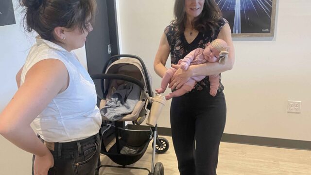 A woman smiles while holding a baby next to a stroller. Another woman stands nearby, facing them. A poster hangs on the wall in the background.