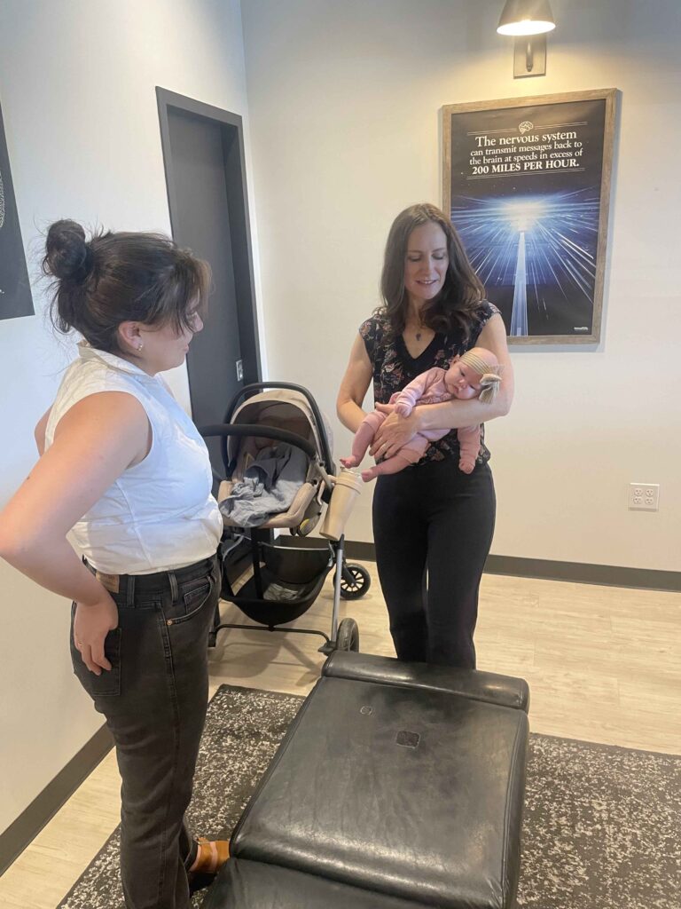 A woman smiles while holding a baby next to a stroller. Another woman stands nearby, facing them. A poster hangs on the wall in the background.