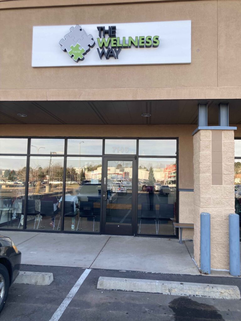 Storefront of "The Wellness Way" with a glass entrance, located in a parking lot. A sign with a puzzle piece logo is displayed above.