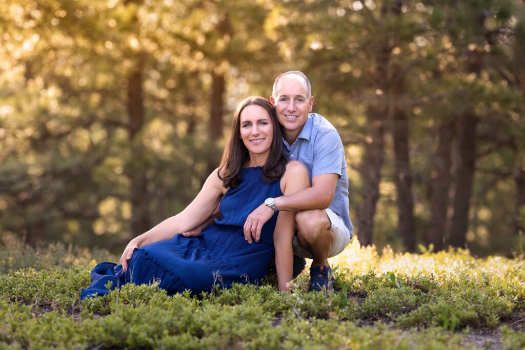 A couple poses outdoors, with the woman sitting and the man kneeling beside her in a forested area with sunlight filtering through trees.