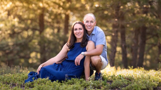 A couple poses outdoors, with the woman sitting and the man kneeling beside her in a forested area with sunlight filtering through trees.