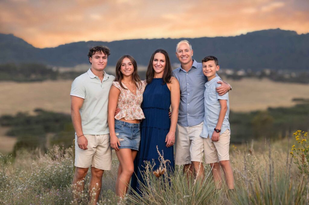 A family of five stands together outdoors in a field at sunset, with hills in the background.