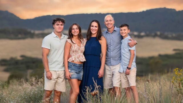 A family of five stands together outdoors in a field at sunset, with hills in the background.