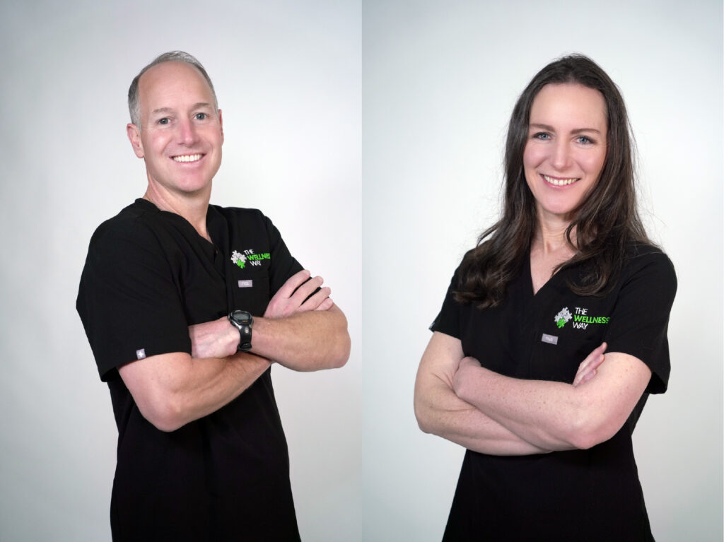 Dr John and Dr Lara Condon of The Wellness Way in black uniforms with "The Wellness Way" logo, standing with arms crossed against a plain background.