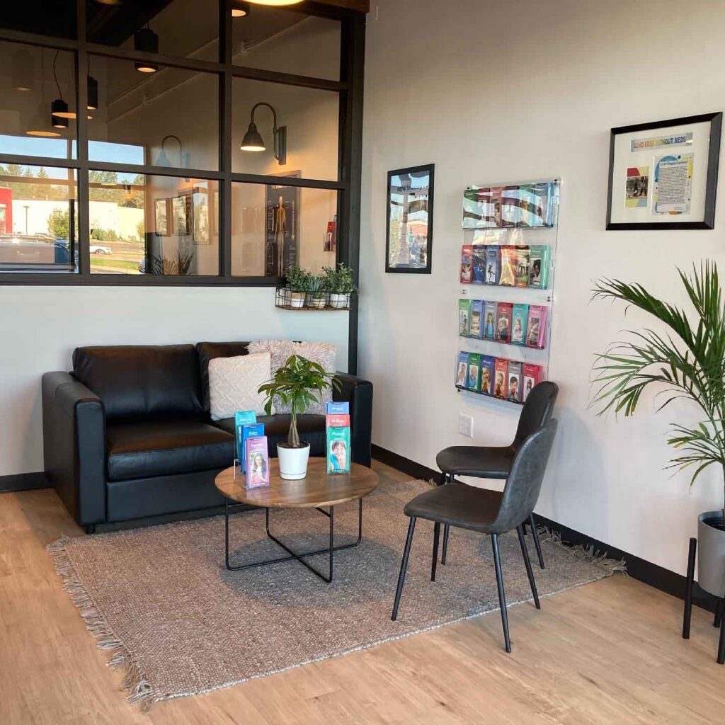 A waiting area with a black sofa, a chair, a round table with plants, brochures on the wall, and framed pictures. The floor is wooden with a textured rug.