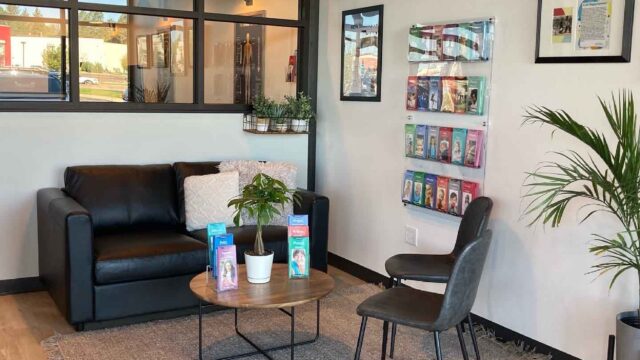 A waiting area with a black sofa, a chair, a round table with plants, brochures on the wall, and framed pictures. The floor is wooden with a textured rug.