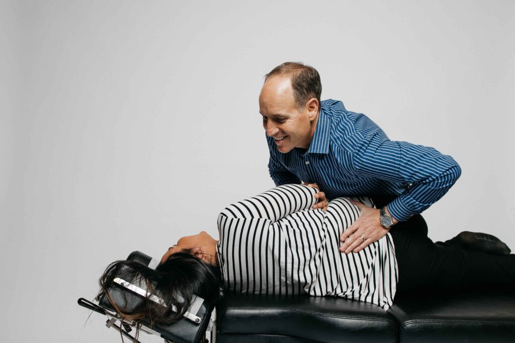 Person in a striped shirt receives a chiropractic adjustment from a person in a blue shirt on a treatment table.