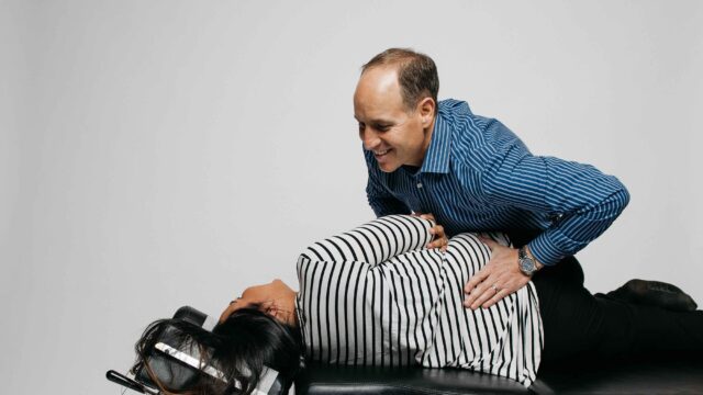 Person in a striped shirt receives a chiropractic adjustment from a person in a blue shirt on a treatment table.