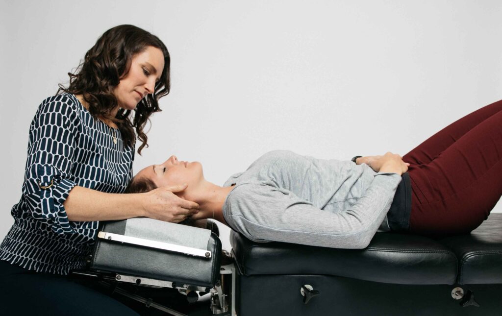 A woman in a patterned blouse performs a chiropractic adjustment on a woman lying down on a treatment table.