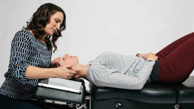 A woman in a patterned blouse performs a chiropractic adjustment on a woman lying down on a treatment table.