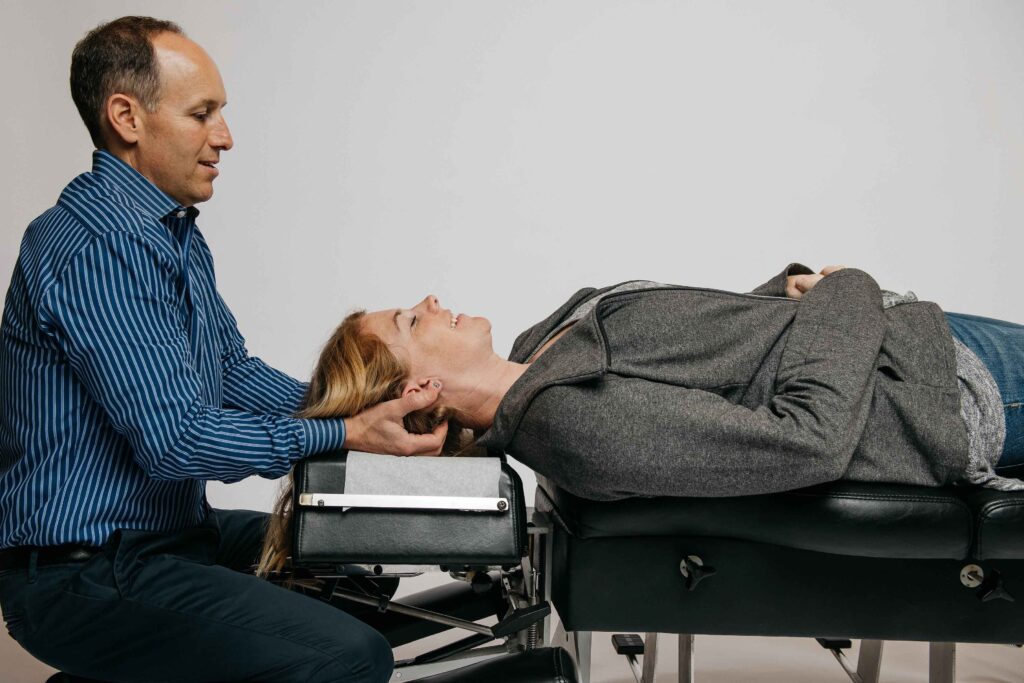 A person lies on a chiropractic table while another person, in a striped shirt, supports their head with both hands during a treatment session.