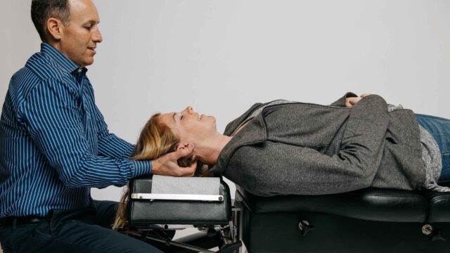 A person lies on a chiropractic table while another person, in a striped shirt, supports their head with both hands during a treatment session.