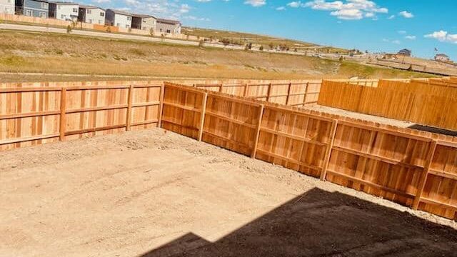 A fenced backyard with bare dirt ground, bordered by wooden privacy fences, with houses and blue sky visible in the background.