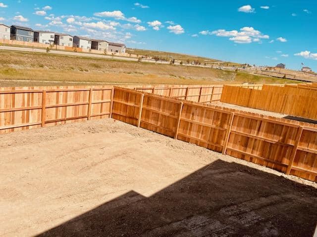 A fenced backyard with bare dirt ground, bordered by wooden privacy fences, with houses and blue sky visible in the background.