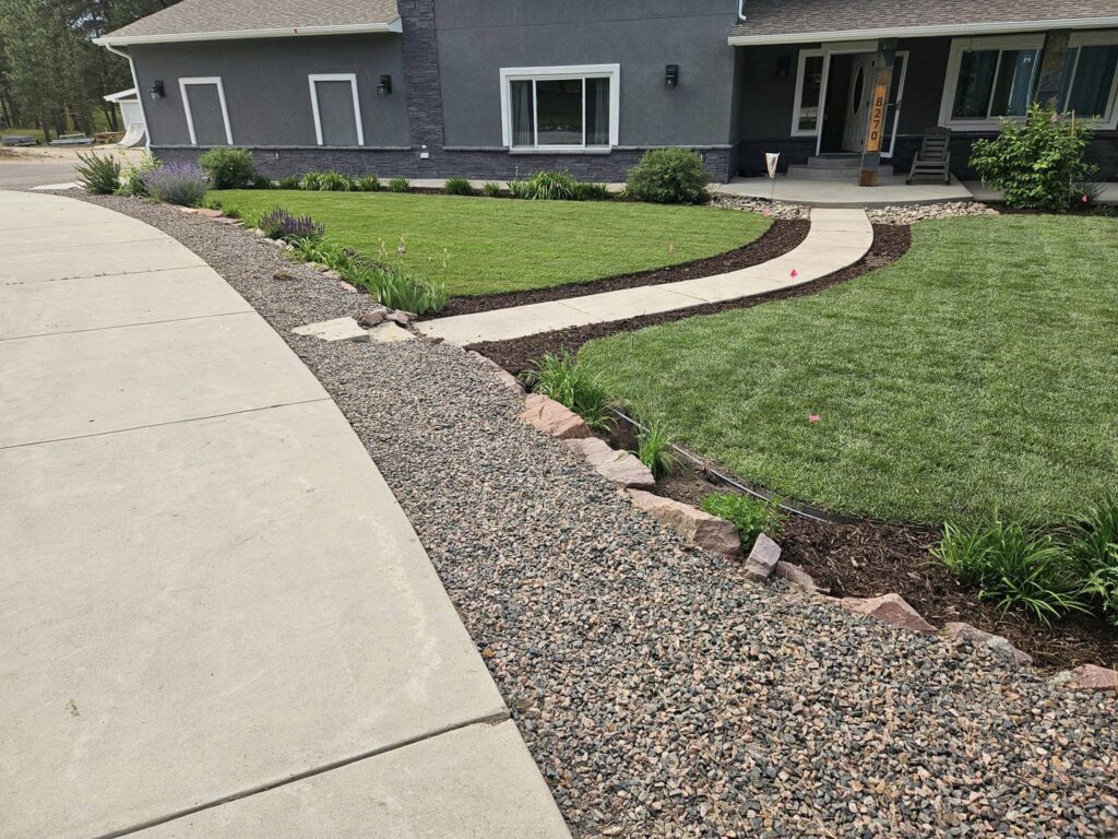 A curved concrete walkway leads through a landscaped front yard with green grass, mulch, small plants, and decorative rock edging near a gray house.
