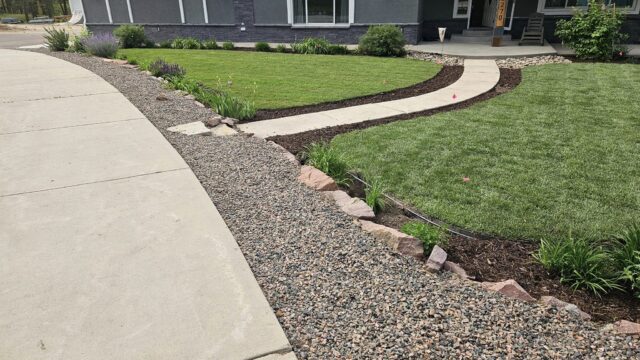 A curved concrete walkway leads through a landscaped front yard with green grass, mulch, small plants, and decorative rock edging near a gray house.