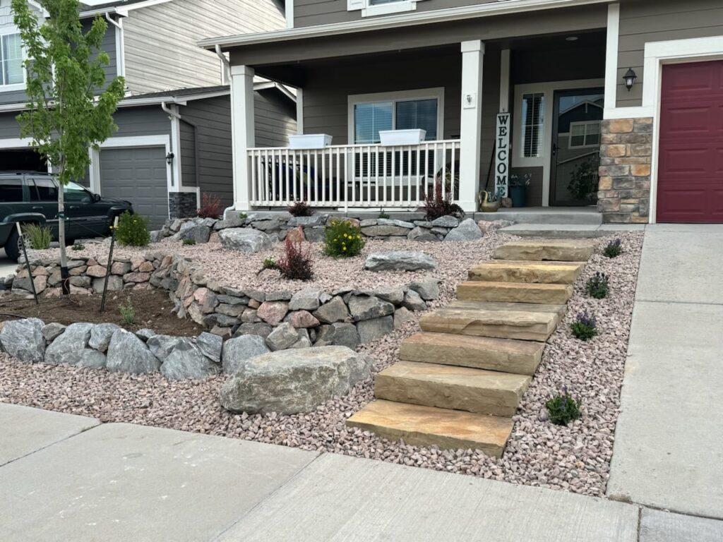 Front yard with stone steps, rock landscaping, small shrubs, and a covered porch with a "Welcome" sign. House exterior is beige with white trim and red garage door.