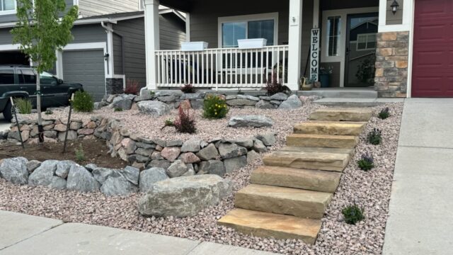 Front yard with stone steps, rock landscaping, small shrubs, and a covered porch with a "Welcome" sign. House exterior is beige with white trim and red garage door.