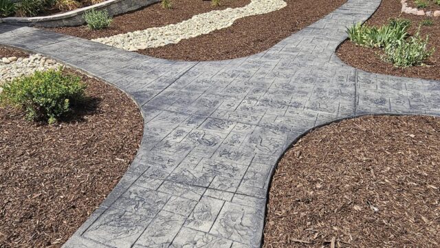 A landscaped yard with a stamped concrete walkway, mulched garden beds, rocks, shrubs, and a small greenhouse in the background near trees.