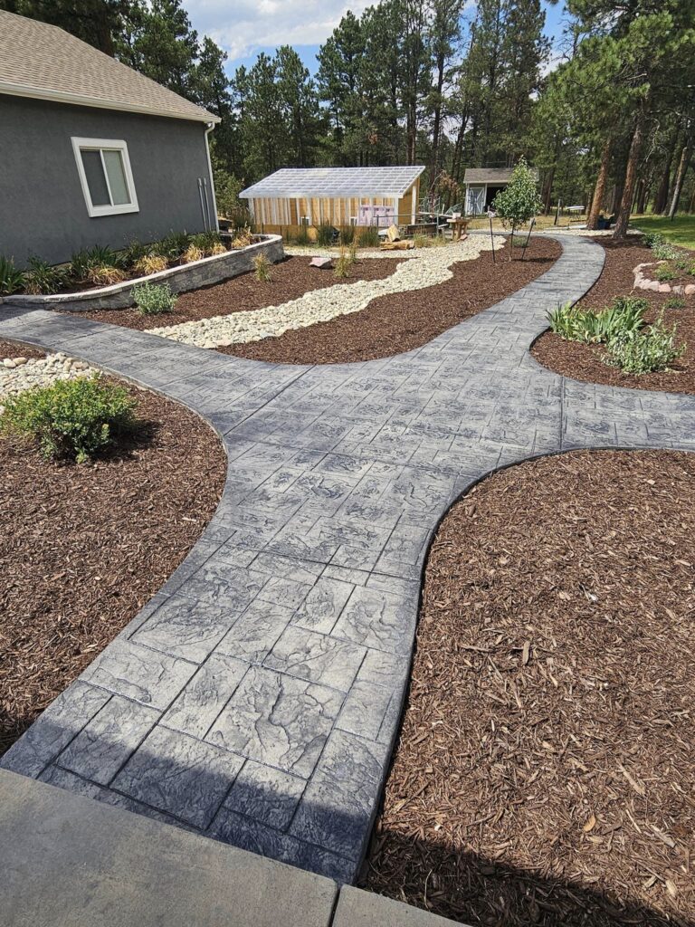 A landscaped yard with a stamped concrete walkway, mulched garden beds, rocks, shrubs, and a small greenhouse in the background near trees.