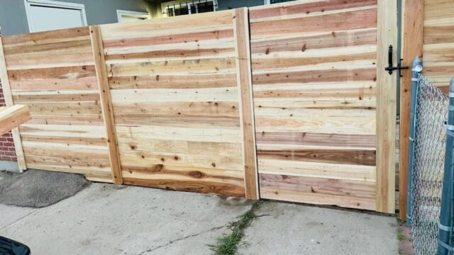 A new wooden privacy fence with a gate stands on a concrete driveway, bordered by a metal chain-link fence and a house in the background.