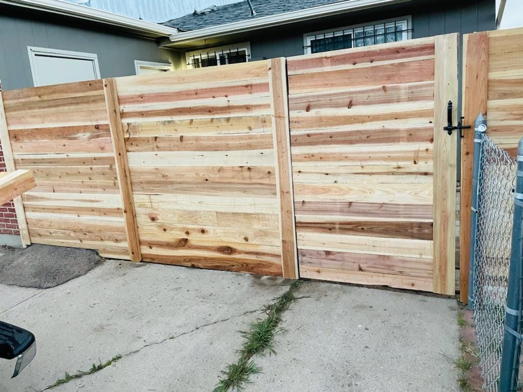 A new wooden privacy fence with a gate stands on a concrete driveway, bordered by a metal chain-link fence and a house in the background.