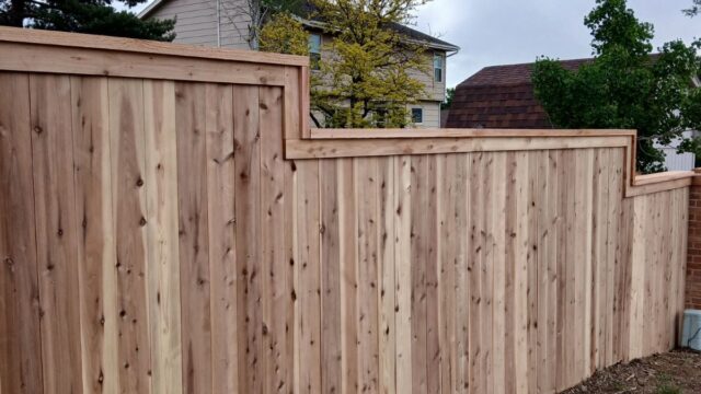 A newly constructed wooden privacy fence with a stepped top design runs alongside a dirt path, with houses and trees in the background.